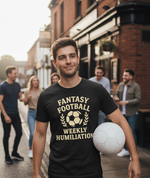 Man wearing a black t-shirt with 'Fantasy Football Weekly Humiliation' text, holding a soccer ball on a street. a Cows Corner t-shirt with the funny football slogan 'Fantasy Football = Weekly Humiliation'. Cows Corner gifts are perfect for sport-mad fans, these gifts work brilliantly for birthdays, new baby celebrations, Father’s Day, Mother’s Day, Christmas, anniversaries, thank you gifts, end-of-season team awards, graduations, retirements, and just-because moments when you want to raise a smile. 
