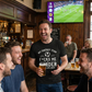 A group of men in a pub watching sports on TV and drinking beer with one man wearing a Cows Corner funny slogan t-shirt