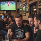 Three men are sitting at a bar watching sports on TV, wearing a Cows Corner funny slogan t-shirt