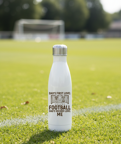 White water bottle with text on a grassy field. This is a Cows Corner water bottle with a funny football slogan 'Dad’s First Love: Football. Dad’s Second Love: Me'. Cows Corner gifts are perfect for sport-mad fans, these gifts work brilliantly for birthdays, new baby celebrations, Father’s Day, Mother’s Day, Christmas, anniversaries, thank you gifts, end-of-season team awards, graduations, retirements, and just-because moments when you want to raise a smile. 
