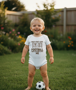 Child wearing a onesie with text in a garden. The baby is wearing a Cows Corner baby grow with the funny football slogan 'Dad’s First Love: Football. Dad’s Second Love: Me'. Cows Corner gifts are perfect for sport-mad fans, these gifts work brilliantly for birthdays, new baby celebrations, Father’s Day, Mother’s Day, Christmas, anniversaries, thank you gifts, end-of-season team awards, graduations, retirements, and just-because moments when you want to raise a smile. 
