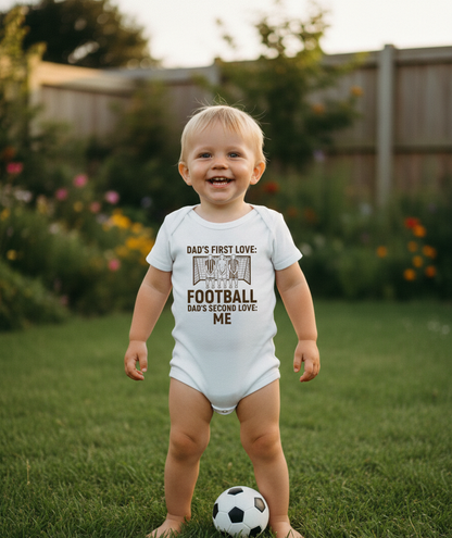Child wearing a onesie with text in a garden. The baby is wearing a Cows Corner baby grow with the funny football slogan 'Dad’s First Love: Football. Dad’s Second Love: Me'. Cows Corner gifts are perfect for sport-mad fans, these gifts work brilliantly for birthdays, new baby celebrations, Father’s Day, Mother’s Day, Christmas, anniversaries, thank you gifts, end-of-season team awards, graduations, retirements, and just-because moments when you want to raise a smile. 
