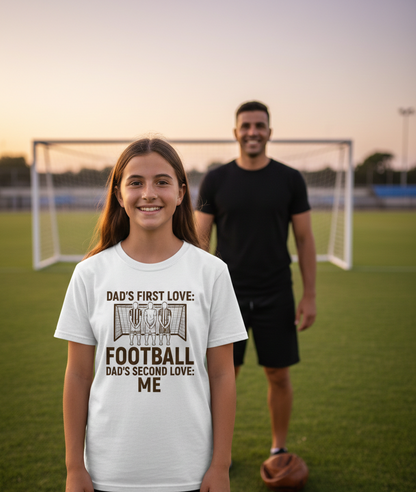 Girl wearing a white t-shirt with a football-themed design on a soccer field with a man in the background. The girl is wearing a Cows Corner t-shirt with the funny football slogan 'Dad’s First Love: Football. Dad’s Second Love: Me'. Cows Corner gifts are perfect for sport-mad fans, these gifts work brilliantly for birthdays, new baby celebrations, Father’s Day, Mother’s Day, Christmas, anniversaries, thank you gifts, end-of-season team awards, graduations, retirements, and just-because moments 