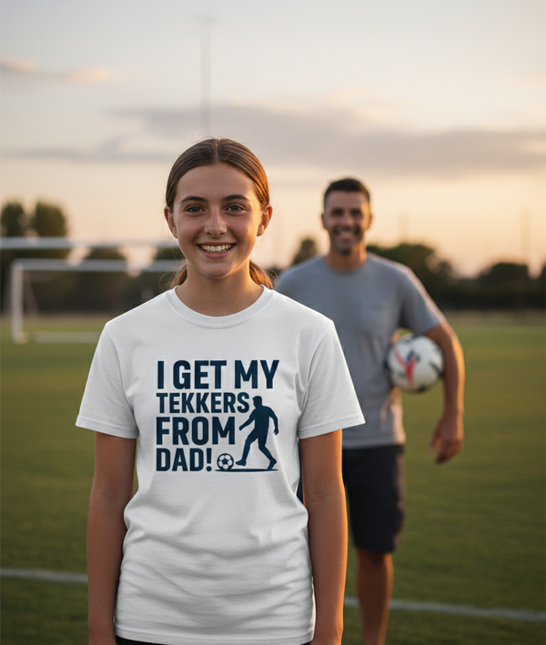 Girl wearing a t-shirt with a soccer-themed message on a sports field with a man in the background. The girl is wearing a Cows Corner t-shirt with the funny football slogan 'I Get My Tekkers From Dad'. Cows Corner gifts are perfect for sport-mad fans, these gifts work brilliantly for birthdays, new baby celebrations, Father’s Day, Mother’s Day, Christmas, anniversaries, thank you gifts, end-of-season team awards, graduations, retirements, and just-because moments when you want to raise a smile. 
