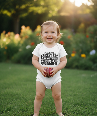 Child wearing a white onesie with text, holding a cricket ball in a garden setting. The baby is wearing a Cows Corner baby grow with a funny cricket slogan 'Dads like an old cricket bat, full of stories but still in the game!'.  Cows Corner gifts are perfect for sport-mad fans, these gifts work brilliantly for birthdays, new baby celebrations, Father’s Day, Mother’s Day, Christmas, anniversaries, thank you gifts, end-of-season team awards, graduations, retirements, and just-because moments 