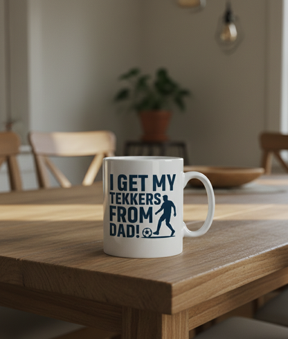 Mug with text on a wooden table in a room with chairs and a plant. This is a Cows Corner mug with a funny football slogan 'I Get My Tekkers From Dad'. Cows Corner gifts are perfect for sport-mad fans, these gifts work brilliantly for birthdays, new baby celebrations, Father’s Day, Mother’s Day, Christmas, anniversaries, thank you gifts, end-of-season team awards, graduations, retirements, and just-because moments when you want to raise a smile. 

