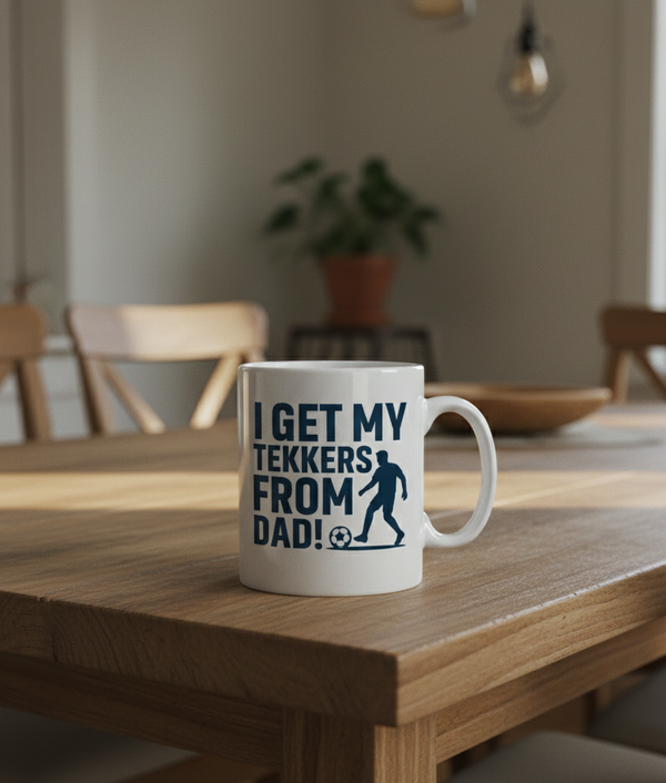 Mug with text on a wooden table in a room with chairs and a plant. This is a Cows Corner mug with a funny football slogan 'I Get My Tekkers From Dad'. Cows Corner gifts are perfect for sport-mad fans, these gifts work brilliantly for birthdays, new baby celebrations, Father’s Day, Mother’s Day, Christmas, anniversaries, thank you gifts, end-of-season team awards, graduations, retirements, and just-because moments when you want to raise a smile. 
