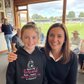 Woman and young girl posing together with a hoodie featuring text in a room with a view of a sports field. The girl is wearing a Cows Corner hoodie with the funny cricket slogan that reads 'Mum's My Personal Third Umpire, Always Reviewing My Choices'.  Cows Corner gifts are perfect for sport-mad fans, these gifts work brilliantly for birthdays, new baby celebrations, Father’s Day, Mother’s Day, Christmas, anniversaries, thank you gifts, end-of-season team awards, graduations, retirements, and just-because