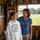 Woman and young girl standing together in a room with sports equipment, smiling. The girl is wearing a Cows Corner hoodie with the funny cricket slogan 'Mums Like A Good Bat Grip, Always Holding Things Together'. Cows Corner gifts are perfect for sport-mad fans, these gifts work brilliantly for birthdays, new baby celebrations, Father’s Day, Mother’s Day, Christmas, anniversaries, thank you gifts, end-of-season team awards, graduations, retirements, and just-because moments when you want to raise a smile. 
