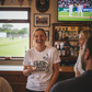 People in a pub watching cricket on TV with a woman holding a drink.  The lady is wearing a Cows Corner T-shirt with the funny cricket slogan on the front that reads 'Club Cricket where Everyone’s an All-Rounder'.  Cows Corner gifts are perfect for sport-mad fans, these gifts work brilliantly for birthdays, new baby celebrations, Father’s Day, Mother’s Day, Christmas, anniversaries, thank you gifts, end-of-season team awards, graduations, retirements, and just-because moments when you want to raise a smile.