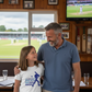 Man and young girl standing together in a pub watching cricket on television. The young girl is wearing a Cows Corner T-shirt with a funny and sweet cricket slogan on the front that reads 'Dad Bowled Me Over With His Dad Jokes'.  Cows Corner gifts are perfect for sport-mad fans who love a bit of humour and personality in their presents. They’re ideal for friends, partners, parents, coaches, teammates, and proud mums and dads who live and breathe sport. 