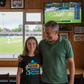 Man and young girl standing together in a room with a TV showing a cricket match.  The young girl is wearing a Cows Corner t-shirt with the funny and sweet cricket slogan on the front that reads 'Dad is a Great Fielder, Always Catching Me Out'