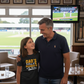 Man and young girl standing together in a room with a TV showing a cricket match.  The young girl is wearing a Cows Corner t-shirt with the funny cricket slogan on the front that reads 'Dads Batting At Number 11 In The Kitchen'