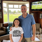 Man and young girl standing together in a room with a TV showing a cricket match with the girl wearing a Cows Corner t-shirt with the funny slogan on the front saying 'Dads like an old cricket bat, full of stories but still in the game!'  Cows Corner gifts are perfect for sport-mad fans who love a bit of humour and personality in their presents. They’re ideal for friends, partners, parents, coaches, teammates, and proud mums and dads who live and breathe sport, whether they play, watch from the sidelines