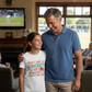 Man and young girl standing together in a living room watching cricket on TV.  The young girl is wearing a Cows Corner t-shirt with the funny and sweet cricket saying on the front that reads 'Dads like a cricket helmet always protecting me'