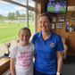 Two women standing in a room with a view of a sports field, smiling at the camera.  The young girl is wearing a Cows Corner t-shirt that has the funny and sweet cricket slogan on the front that reads 'Mums Like A Good All-rounder, Does Everything'