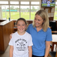 Woman and young girl standing together in a sports facility with a TV screen in the background.  The young girl is wearing a Cows Corner t-shirt with the funny and sweet cricket saying on the front that reads 'Mums Like A Good Bat Grip, Always Holding Things Together'