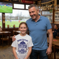 Man and young girl standing together in a sports bar with a TV showing a football match.  The girl is wearing a Cows Corner t-shirt with the funny and sweet football slogan on the front that reads 'Born To Play Football. Raised by a Football Dad!'. Cows Corner gifts are perfect for sport-mad fans, these gifts work brilliantly for birthdays, new baby celebrations, Father’s Day, Mother’s Day, Christmas, anniversaries, thank you gifts, end-of-season team awards, graduations, retirements, and just-because