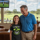 Man and young girl standing together in a golf-themed room with a TV screen showing a golf game. The girl is wearing a Cows Corner t-shirt with a funny and sweet golf saying on the front that reads 'My Dad’s Golf Game Is Legendary in His Own Mind!'. Cows Corner gifts are perfect for sport-mad fans, these gifts work brilliantly for birthdays, new baby celebrations, Father’s Day, Mother’s Day, Christmas, anniversaries, thank you gifts, end-of-season team awards, graduations, retirements, and just-because 
