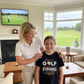 Woman and young girl in a living room with a TV showing golf, a fireplace, and a window view of a golf course. The girl is wearing a Cows Corner t-shirt with the funny and sweet golf saying that reads on the front 'Mum’s Golf Game Is As Strong as Her Coffee!'. Cows Corner gifts are perfect for sport-mad fans, these gifts work brilliantly for birthdays, new baby celebrations, Father’s Day, Mother’s Day, Christmas, anniversaries, thank you gifts, end-of-season team awards, graduations, retirements