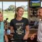 Woman wearing a black t-shirt with a humorous golf quote, holding a glass of wine, in a golf-themed bar.  The lady is wearing a Cows Corner t-shirt with a funny golf slogan on the front that reads 'Your Golf Game Bring Me Joy, Because It’s Worse Than Mine'. Cows Corner gifts are perfect for sport-mad fans, these gifts work brilliantly for birthdays, new baby celebrations, Father’s Day, Mother’s Day, Christmas, anniversaries, thank you gifts, end-of-season team awards, graduations, retirements, and just-beca