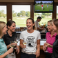 Group of women in a bar with golf-themed decor, watching a golf game on TV. The lady is wearing a Cows Corner T-shirt with the funny golf slogan on the front that reads 'Your Swing Is Proof That Money Can’t Buy Skill'. Cows Corner gifts are perfect for sport-mad fans, these gifts work brilliantly for birthdays, new baby celebrations, Father’s Day, Mother’s Day, Christmas, anniversaries, thank you gifts, end-of-season team awards, graduations, retirements, and just-because moments 