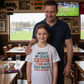 Man and young girl standing together in a sports bar with a TV showing a game. The girl is wearing a Cows Corner t-shirt with the funny and sweet rugby slogan on the front that reads 'Dad’s My Coach My Biggest Fan and My Post-Match Snack Provider!'. Cows Corner gifts are perfect for sport-mad fans, these gifts work brilliantly for birthdays, new baby celebrations, Father’s Day, Mother’s Day, Christmas, anniversaries, thank you gifts, end-of-season team awards, graduations, retirements, and just-because
