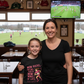 Woman and young girl in a sports bar with a TV showing a soccer match. The girl is wearing a Cows Corner t-shirt with the funny and sweet slogan that reads 'Mum Rucks, Mum Tackles, Mum Wins at Life!'. Cows Corner gifts are perfect for sport-mad fans, these gifts work brilliantly for birthdays, new baby celebrations, Father’s Day, Mother’s Day, Christmas, anniversaries, thank you gifts, end-of-season team awards, graduations, retirements, and just-because moments when you want to raise a smile. 
