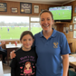 Woman and young girl standing together in a sports bar with a TV showing a soccer match. The girl is wearing a Cows Corner t-shirt with a funny and sweet rugby slogan that reads 'Mum’s My Toughest Coach and My Softest Hug!'. Cows Corner gifts are perfect for sport-mad fans, these gifts work brilliantly for birthdays, new baby celebrations, Father’s Day, Mother’s Day, Christmas, anniversaries, thank you gifts, end-of-season team awards, graduations, retirements, and just-because moments 