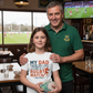 Man and young girl in a pub with a rugby ball, wearing a shirt with a humorous rugby quote. The girl is wearing a Cows Corner t-shirt with a funny rugby slogan on the front that reads 'My Dad Tackles Life Like a Rugby Match- Head First'. Cows Corner gifts are perfect for sport-mad fans, these gifts work brilliantly for birthdays, new baby celebrations, Father’s Day, Mother’s Day, Christmas, anniversaries, thank you gifts, end-of-season team awards, graduations, retirements, and just-because moments 
