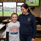 Woman and young girl standing together in a sports-themed room with a TV showing a rugby match. Girl wearing Cows Corner t-shirt with the funny and sweet rugby slogan that reads 'My Mum Tackles Everything on the Pitch and at Home!' . Cows Corner gifts are perfect for sport-mad fans, these gifts work brilliantly for birthdays, new baby celebrations, Father’s Day, Mother’s Day, Christmas, anniversaries, thank you gifts, end-of-season team awards, graduations, retirements, and just-because moments 