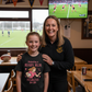 Woman and young girl in a sports bar with rugby-themed decor and TV showing a rugby match. The girl is wearing a Cows Corner t-shirt with the funny and sweet rugby slogan 'Mum Rucks, Mum Tackles, Mum Wins at Life!'. Cows Corner gifts are perfect for sport-mad fans, these gifts work brilliantly for birthdays, new baby celebrations, Father’s Day, Mother’s Day, Christmas, anniversaries, thank you gifts, end-of-season team awards, graduations, retirements, and just-because moments when you want to raise a smile