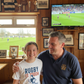 Man and young girl in a room with sports memorabilia and a TV showing a rugby match.  The girl is wearing a Cows Corner t-shirt with the funny rugby slogan on the front that reads 'Rugby Is Tough but My Dad Is Tougher!'  Cows Corner gifts are perfect for sport-mad fans, these gifts work brilliantly for birthdays, new baby celebrations, Father’s Day, Mother’s Day, Christmas, anniversaries, thank you gifts, end-of-season team awards, graduations, retirements, and just-because moments 