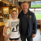 Two people standing in a pub with a television showing a sports match in the background. Girl wearing a Cows Corner t-shirt with the rugby coach slogan on the front that reads 'You Taught Me More Than Rugby, You Taught Me Resilience. Thank You, Coach'. Cows Corner gifts are perfect for sport-mad fans, these gifts work brilliantly for birthdays, new baby celebrations, Father’s Day, Mother’s Day, Christmas, anniversaries, thank you gifts, end-of-season team awards, graduations, retirements, and just-because