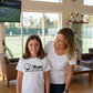 Woman and young girl standing in a living room with a tennis court view, both wearing white t-shirts.  The girl is wearing a Cows Corner T-shirt with the funny and sweet tennis saying on the front that reads 'Mum Serves Up Love and Aces'. Cows Corner gifts are perfect for sport-mad fans, these gifts work brilliantly for birthdays, new baby celebrations, Father’s Day, Mother’s Day, Christmas, anniversaries, thank you gifts, end-of-season team awards, graduations, retirements, and just-because moments 
