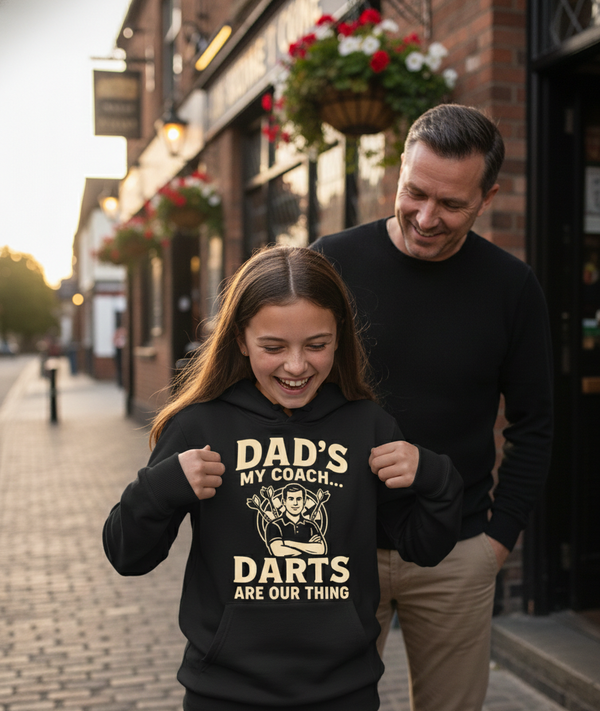 A girl wearing a Cows Corner hoodie with the funny darts slogan 'Dad’s My Coach… Darts Are Our Thing'. Cows Corner gifts are perfect for sport-mad fans, these gifts work brilliantly for birthdays, new baby celebrations, Father’s Day, Mother’s Day, Christmas, anniversaries, thank you gifts, end-of-season team awards, graduations, retirements, and just-because moments when you want to raise a smile. 

