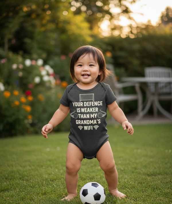 Child wearing a onesie with text standing on grass with a soccer ball. The baby is wearing a Cows Corner baby grow with a funny football slogan 'Your Defence Is Weaker Than My Grandma’s Wifi'. Cows Corner gifts are perfect for sport-mad fans, these gifts work brilliantly for birthdays, new baby celebrations, Father’s Day, Mother’s Day, Christmas, anniversaries, thank you gifts, end-of-season team awards, graduations, retirements, and just-because moments when you want to raise a smile. 
