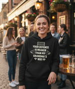 Person wearing a black hoodie with humorous text in an outdoor pub setting. The lady is wearing a Cows Corner hoodie with a funny football slogan 'Your Defence Is Weaker Than My Grandma’s Wifi'. Cows Corner gifts are perfect for sport-mad fans, these gifts work brilliantly for birthdays, new baby celebrations, Father’s Day, Mother’s Day, Christmas, anniversaries, thank you gifts, end-of-season team awards, graduations, retirements, and just-because moments when you want to raise a smile. 
