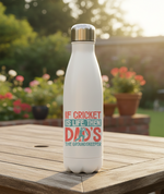 White water bottle with a colorful text design on a wooden table outdoors. This is a Cows Corner water bottle with a funny cricket slogan 'If Cricket Is Life, Then Dad's The Groundkeeper'. Cows Corner gifts are perfect for sport-mad fans, these gifts work brilliantly for birthdays, new baby celebrations, Father’s Day, Mother’s Day, Christmas, anniversaries, thank you gifts, end-of-season team awards, graduations, retirements, and just-because moments when you want to raise a smile. 
