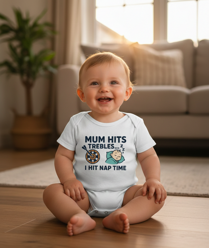 Baby wearing a white onesie with humorous text in a living room. The baby is wearing a Cows Corner baby grow with a funny darts slogan 'Mum Hits Trebles… I Hit Nap Time'. Cows Corner gifts are perfect for sport-mad fans, these gifts work brilliantly for birthdays, new baby celebrations, Father’s Day, Mother’s Day, Christmas, anniversaries, thank you gifts, end-of-season team awards, graduations, retirements, and just-because moments when you want to raise a smile. 
