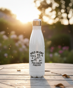 White water bottle with a printed design on a wooden surface with a blurred natural background. This is a Cows Corner water bottle with a funny golf slogan 'Hole-In-One Today… Pub Stories Forever'. Cows Corner gifts are perfect for sport-mad fans, these gifts work brilliantly for birthdays, new baby celebrations, Father’s Day, Mother’s Day, Christmas, anniversaries, thank you gifts, end-of-season team awards, graduations, retirements, and just-because moments when you want to raise a smile. 
