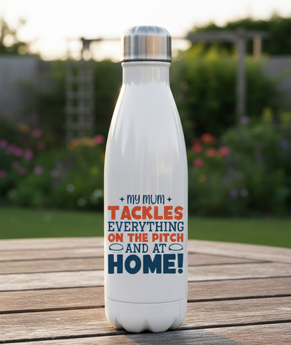 White water bottle with colorful text on a wooden surface with a garden background. This is a Cows Corner water bottle with a funny rugby slogan 'My Mum Tackles Everything on the Pitch and at Home!'. Cows Corner gifts are perfect for sport-mad fans, these gifts work brilliantly for birthdays, new baby celebrations, Father’s Day, Mother’s Day, Christmas, anniversaries, thank you gifts, end-of-season team awards, graduations, retirements, and just-because moments when you want to raise a smile. 
