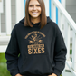 A lady wearing a black hoodie with You Bring the Sledging, I'll Bring the Sixes on the front. A Funny Cricket Hoodie, Premium Fabric, Multiple Adult Sizes