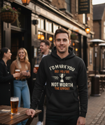 Man wearing a hoodie with a humorous message in an outdoor pub setting. The man is wearing a Cows Corner hoodie with a funny football slogan 'I’d Mark You but You’re Not Worth the Effort'. Cows Corner gifts are perfect for sport-mad fans, these gifts work brilliantly for birthdays, new baby celebrations, Father’s Day, Mother’s Day, Christmas, anniversaries, thank you gifts, end-of-season team awards, graduations, retirements, and just-because moments when you want to raise a smile. 
