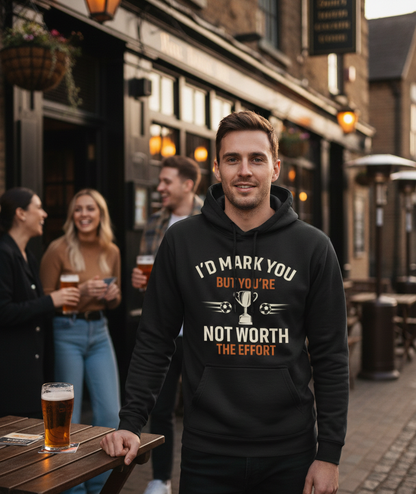 Man wearing a hoodie with a humorous message in an outdoor pub setting. The man is wearing a Cows Corner hoodie with a funny football slogan 'I’d Mark You but You’re Not Worth the Effort'. Cows Corner gifts are perfect for sport-mad fans, these gifts work brilliantly for birthdays, new baby celebrations, Father’s Day, Mother’s Day, Christmas, anniversaries, thank you gifts, end-of-season team awards, graduations, retirements, and just-because moments when you want to raise a smile. 
