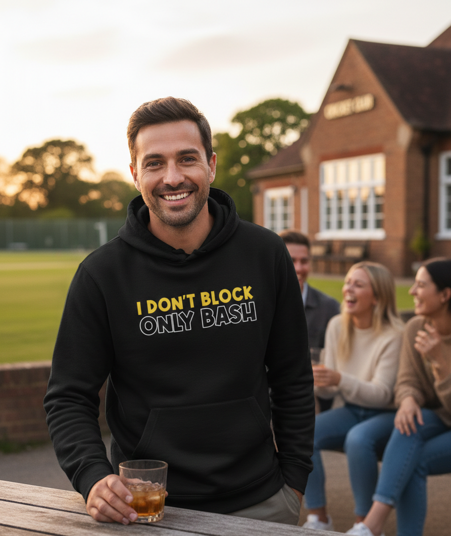 Man wearing a black hoodie with humorous text, sitting outdoors with friends. The man is wearing a Cows Corner hoodie with a funny cricket slogan 'I Don't Block, Only Bash'. Cows Corner gifts are perfect for sport-mad fans, these gifts work brilliantly for birthdays, new baby celebrations, Father’s Day, Mother’s Day, Christmas, anniversaries, thank you gifts, end-of-season team awards, graduations, retirements, and just-because moments when you want to raise a smile. 
