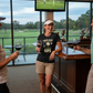 Woman standing in a golf-themed bar with a TV screen showing a golf game. The lady is wearing a Cows Corner t-shirt with a funny golf slogan on the front that reads 'Your Slice Should Come With a GPS'. Cows Corner gifts are perfect for sport-mad fans, these gifts work brilliantly for birthdays, new baby celebrations, Father’s Day, Mother’s Day, Christmas, anniversaries, thank you gifts, end-of-season team awards, graduations, retirements, and just-because moments when you want to raise a smile. 