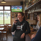 Woman in a pub holding a glass of red wine, smiling. The lady is wearing a Cows Corner hoodie that has the funny cricket slogan 'Bowled & Bamboozled and still looking better than you'. Cows Corner gifts are perfect for sport-mad fans, these gifts work brilliantly for birthdays, new baby celebrations, Father’s Day, Mother’s Day, Christmas, anniversaries, thank you gifts, end-of-season team awards, graduations, retirements, and just-because moments when you want to raise a smile. 

