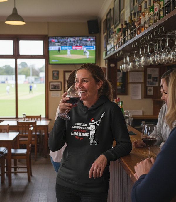 Woman in a pub holding a glass of red wine, smiling. The lady is wearing a Cows Corner hoodie that has the funny cricket slogan 'Bowled & Bamboozled and still looking better than you'. Cows Corner gifts are perfect for sport-mad fans, these gifts work brilliantly for birthdays, new baby celebrations, Father’s Day, Mother’s Day, Christmas, anniversaries, thank you gifts, end-of-season team awards, graduations, retirements, and just-because moments when you want to raise a smile. 

