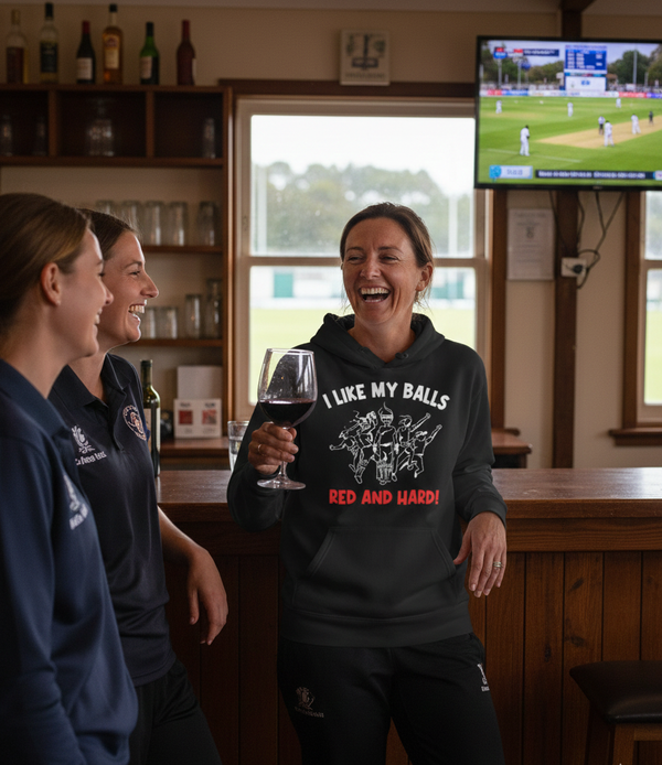 Three women in a pub setting with one holding a glass of wine and wearing a humorous hoodie. The lady is wearing a Cows Corner hoodie with the funny cricket slogan that reads 'I Like My Balls Red And Hard'.  Cows Corner gifts are perfect for sport-mad fans, these gifts work brilliantly for birthdays, new baby celebrations, Father’s Day, Mother’s Day, Christmas, anniversaries, thank you gifts, end-of-season team awards, graduations, retirements, and just-because moments when you want to raise a smile. 

